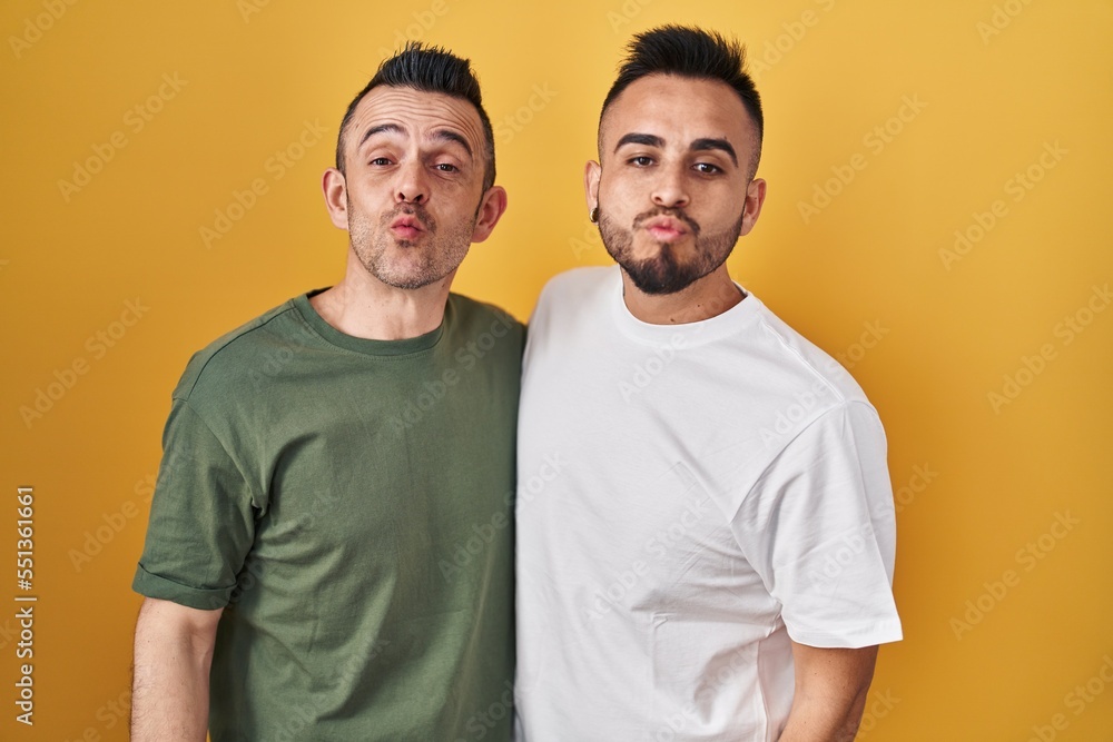 Poster homosexual couple standing over yellow background looking at the camera blowing a kiss on air being 