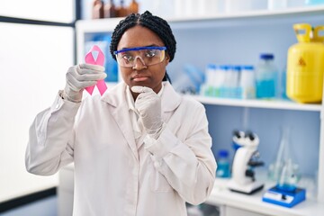 African american woman working at scientist laboratory holding pink ribbon serious face thinking about question with hand on chin, thoughtful about confusing idea