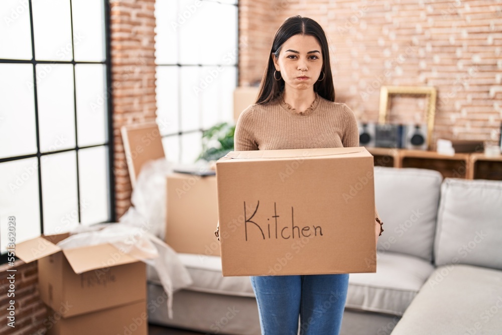 Poster young woman moving to a new home holding box puffing cheeks with funny face. mouth inflated with air