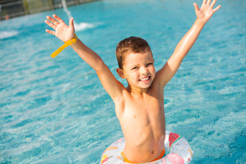 A cheerful child boy in bright turquoise and orange swimming trunks stands with an inflatable ringin the pool with hands up. Child boy having fun in the pool.