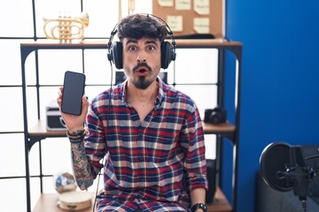 Young hispanic man with beard showing smartphone screen at music studio scared and amazed with open mouth for surprise, disbelief face