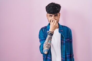 Young hispanic man with beard standing over pink background smelling something stinky and...