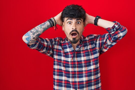Young hispanic man with beard standing over red background crazy and scared with hands on head, afraid and surprised of shock with open mouth