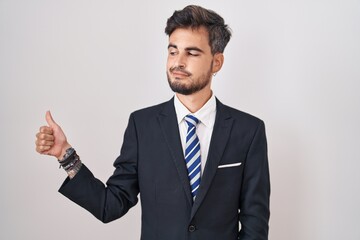 Young hispanic man with tattoos wearing business suit and tie looking proud, smiling doing thumbs up gesture to the side