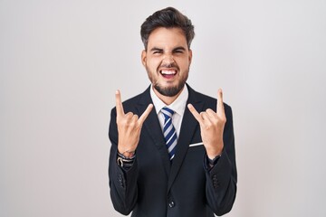 Young hispanic man with tattoos wearing business suit and tie shouting with crazy expression doing rock symbol with hands up. music star. heavy music concept.