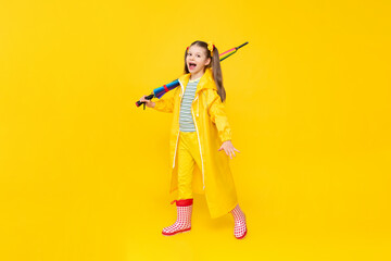 A little girl with a multicolored umbrella in a raincoat and rubber boots on a yellow isolated background. Rainy spring weather and puddles.