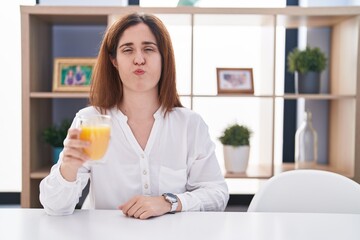 Brunette woman drinking glass of orange juice puffing cheeks with funny face. mouth inflated with air, crazy expression.