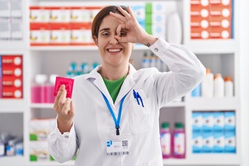 Brunette woman working at pharmacy drugstore holding condom smiling happy doing ok sign with hand...