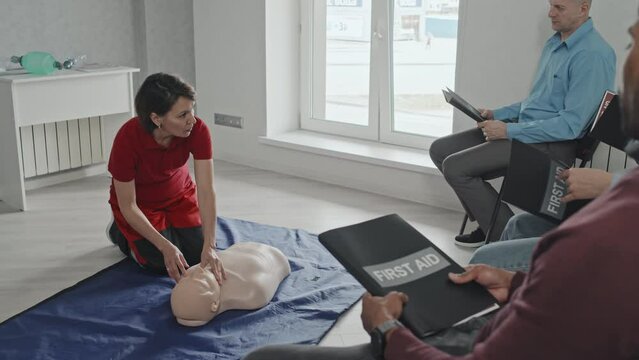 Mature female paramedic in uniform sitting on floor teaching group of people how to perform first aid and CPR