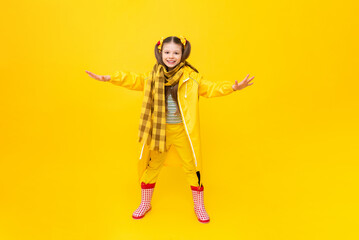 A child in a raincoat and scarf, in rubber red boots on a yellow isolated background. Rainy spring autumn weather. Clothes for the rain.
