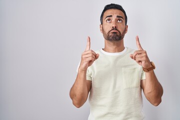 Hispanic man with beard standing over isolated background pointing up looking sad and upset,...