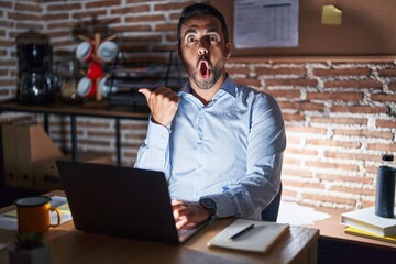 Hispanic man with beard working at the office at night surprised pointing with hand finger to the side, open mouth amazed expression.