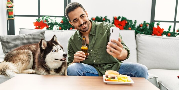 Young hispanic man make selfie sitting on sofa with dog by christmas decor at home