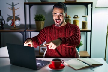 Young hispanic man with beard using computer laptop at night at home pointing to you and the camera with fingers, smiling positive and cheerful