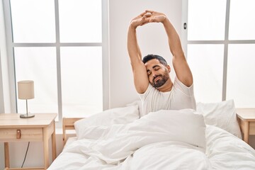 Fototapeta na wymiar Young hispanic man stretching arms sitting on bed at bedroom