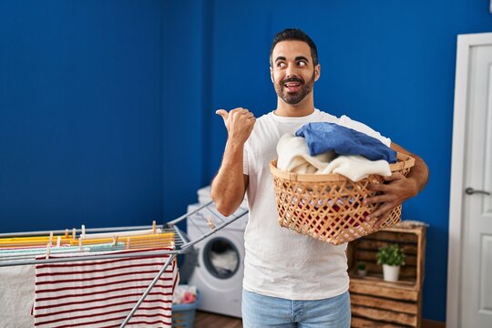 Young hispanic man with beard holding laundry basket pointing thumb up to the side smiling happy with open mouth