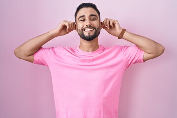 Hispanic young man standing over pink background smiling pulling ears with fingers, funny gesture....