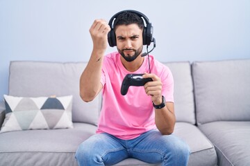 Hispanic young man playing video game holding controller sitting on the sofa doing italian gesture...