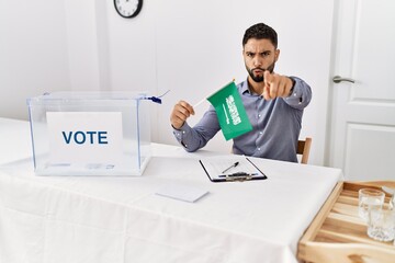 Young handsome man with beard at political campaign election holding arabia saudita flag pointing...