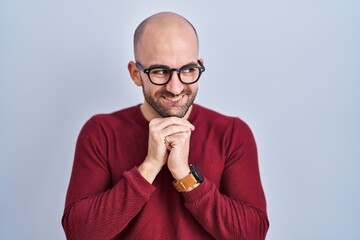 Young bald man with beard standing over white background wearing glasses laughing nervous and excited with hands on chin looking to the side