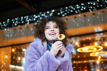 curly haired woman in trendy violet coat drinks mulled wine outdoors, festive background 