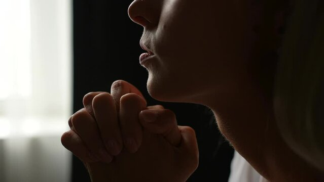 Close-up cropped shot face of unrecognizable sad young woman praying sincerely with folded arms and closed eyes, asking with hopeful imploring expression, begging apology by window in dark room.