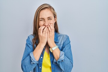 Young blonde woman standing over blue background laughing and embarrassed giggle covering mouth...