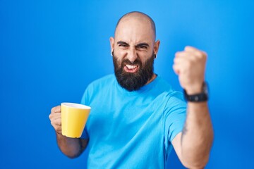 Young hispanic man with beard and tattoos drinking a cup of coffee angry and mad raising fist frustrated and furious while shouting with anger. rage and aggressive concept.