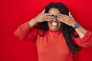 Plus size hispanic woman standing over red background covering eyes with hands smiling cheerful and funny. blind concept.