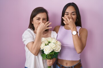 Hispanic mother and daughter holding bouquet of white flowers smelling something stinky and disgusting, intolerable smell, holding breath with fingers on nose. bad smell