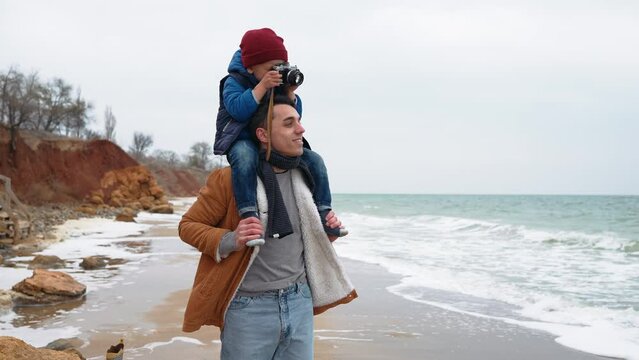 Confident son takes photo on camera on his father neck on the beach near the sea