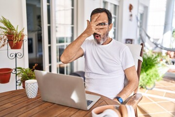 Middle age man using computer laptop at home peeking in shock covering face and eyes with hand,...