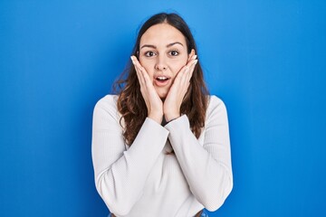 Young hispanic woman standing over blue background afraid and shocked, surprise and amazed expression with hands on face