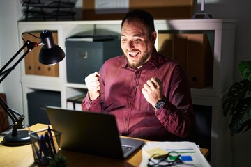 Plus size hispanic man with beard working at the office at night celebrating surprised and amazed for success with arms raised and open eyes. winner concept.