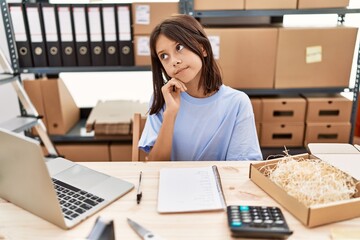 Young hispanic girl working at small business ecommerce serious face thinking about question with...