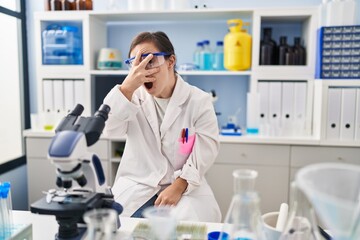 Fototapeta premium Hispanic girl with down syndrome working at scientist laboratory peeking in shock covering face and eyes with hand, looking through fingers with embarrassed expression.