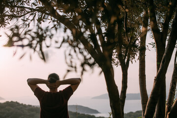 A young man peacefully looks at the sunset with his hands behind his head in a garden overlooking the mountains and the sea.