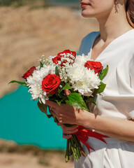 a bouquet of white chrysanthemums and red roses in the hands of the bride against the background of an azure lake.