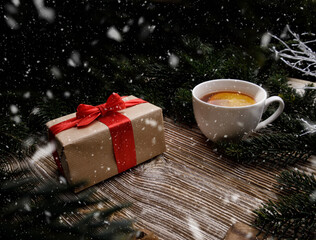 gift box with a red bow and a cup with a fragrant hot drink with fruit stands on a wooden table between fir branches. concept for Christmas and New Year holidays.snow falls