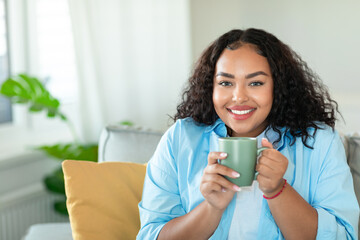 Domestic leisure. Happy black plus size lady drinking coffee at home, resting on comfortable sofa in cozy living room