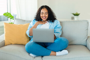 Happy black oversize lady using laptop at home, reading news or making video call with friends,...
