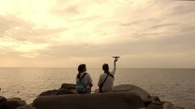 Lovely couple is sitting on the rocky coast in front of the sea enjoying the view and piloting drone filmed in slow motion. Man and woman are resting near the ocean at twilight and flying quadcopter