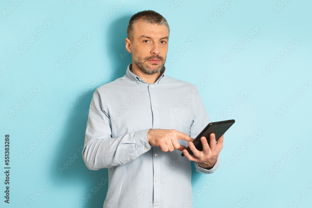Poster serious middle aged caucasian man using tablet computer, standing over blue studio background