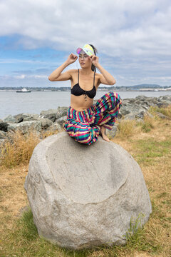 Pretty Asian woman wearing a bikini top and colorful striped pants sits on a rock near the water in Washington state, USA