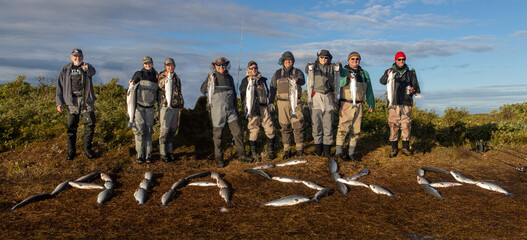 Group of 9 fishermen made up of men and women holding large salmon on a river bank. More fish spell our the word Alaska on the tundra