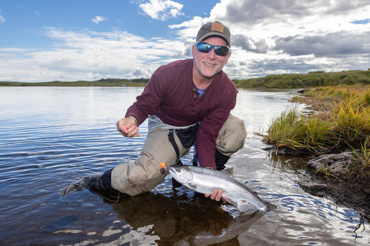 Happy fisherman holding a hooked coho salmon fish above the water of the Egegik river in Alaska