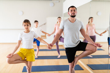 Young girl holding hand of her father while standing in tree pose during family yoga training in fitness studio.