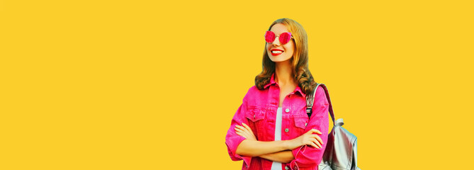 Portrait of beautiful smiling young woman looking away wearing pink jacket, backpack and sunglasses on yellow background, blank copy space for advertising text
