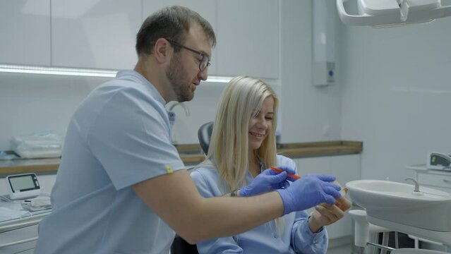 Male dentist explains to female patient how to properly brush her teeth on mock-up. Preventive reception. Slow motion