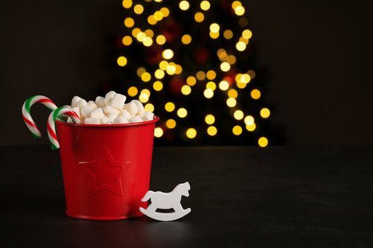 Red bucket with a star full of marshmallows, colorful hard candy, horse decoration, Christmas tree with defocused lights,dark background, place for text